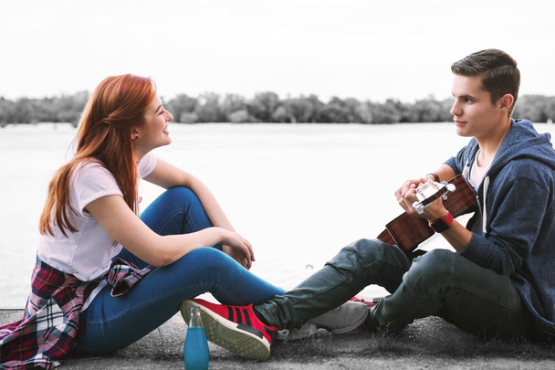 Couple of happy beaming teenagers enjoying nice evening with guitar near the river - Zdjęcie, obraz