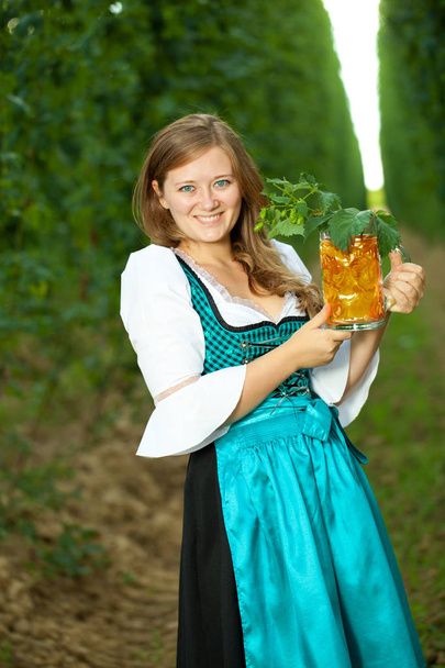 Beautiful young woman in traditional bavarian dress holding mug of beer in the garden. Oktoberfest concept - Fotografie, Obrázek