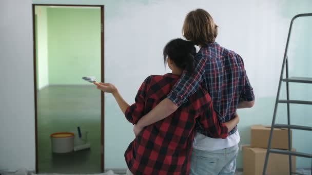 Rear view of renovation diy couple with paintbrush and paint roller embracing and looking at half painted wall in new house. Creative couple discussing idea of new interior design during home makeover - Footage, Video