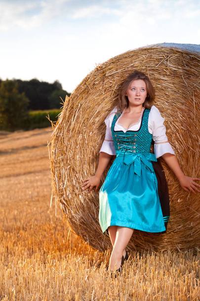 Beautiful young woman in blue dress posing near haystack on wheat field - Fotografie, Obrázek