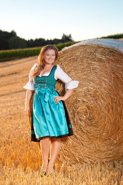 Beautiful young woman in blue dress posing near haystack on wheat field - Fotografie, Obrázek
