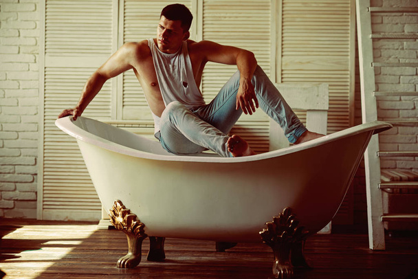 Man in bathroom, hygiene. Sexy macho in tank top and jeans fashion. Guy sit on bath. Fitness model with strong body health. Hygiene and bathing concept - Photo, image