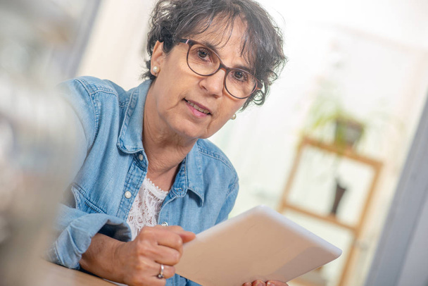 a charming senior brunette woman with glasses using digital tablet at home - Photo, Image
