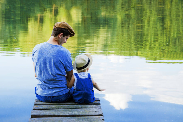 Dad and a tiny girl in dungarees relax at the river. Back view, copy space. - Zdjęcie, obraz