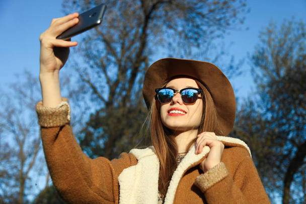Young beautiful caucasian smiling girl in brown hat and sunglasses makes selfie on smartphone in park on sunny day against sky. - Fotó, kép
