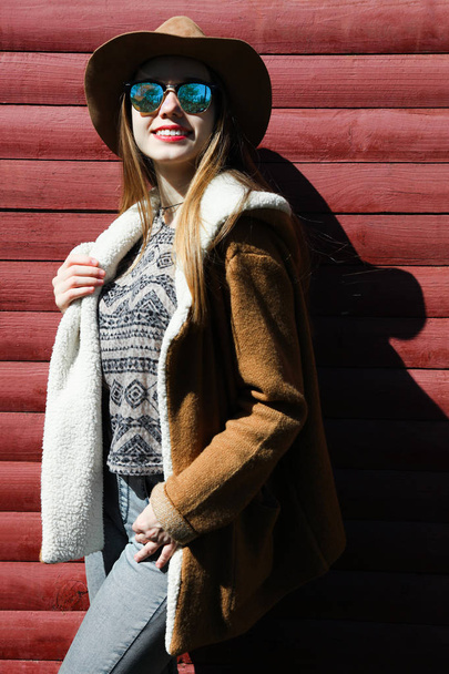 junge schöne kaukasische lächelnde Mädchen mit braunem Hut, Sonnenbrille und Knick auf Hintergrund hölzerne Wand. Landhausstil. - Foto, Bild