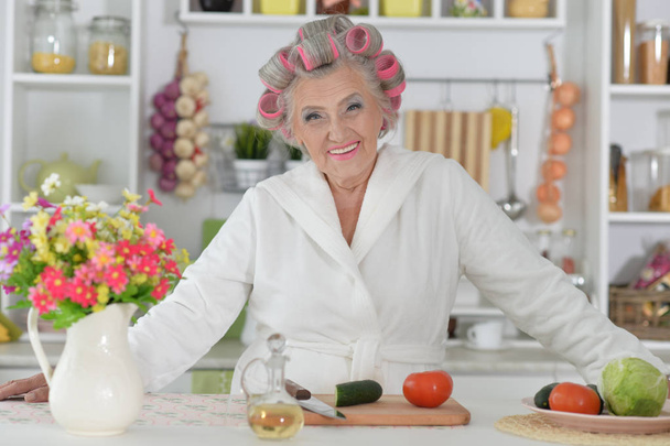 Beautiful senior woman wearing white bathrobe and hair curlers cooking at kitchen - 写真・画像