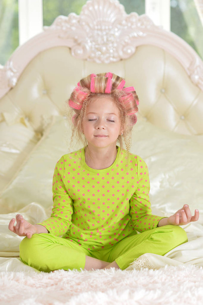 Lovely little girl with pink curlers meditating on bed at home - Fotó, kép