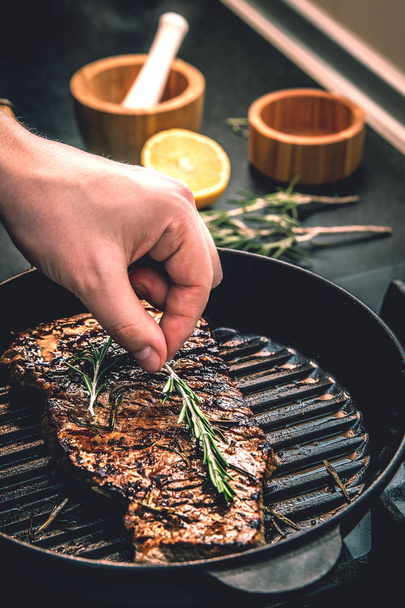 The cook lays a sprig of rosemary on an appetizing juicy beef steak, roasting in a pan for grilling. - Foto, Imagem