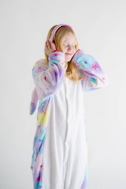 modern fashion - beautiful blonde girl listens to music with headphones on a white background in kigurumi pajamas - Photo, Image