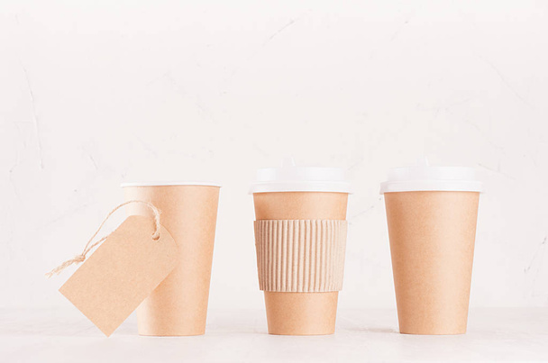 Coffee packing mockup - set of three craft brown paper cups with white caps, cup holder and blank label on white wood board, coffee shop interior. Modern elegant concept for branding identity, advertising, design. - Photo, Image