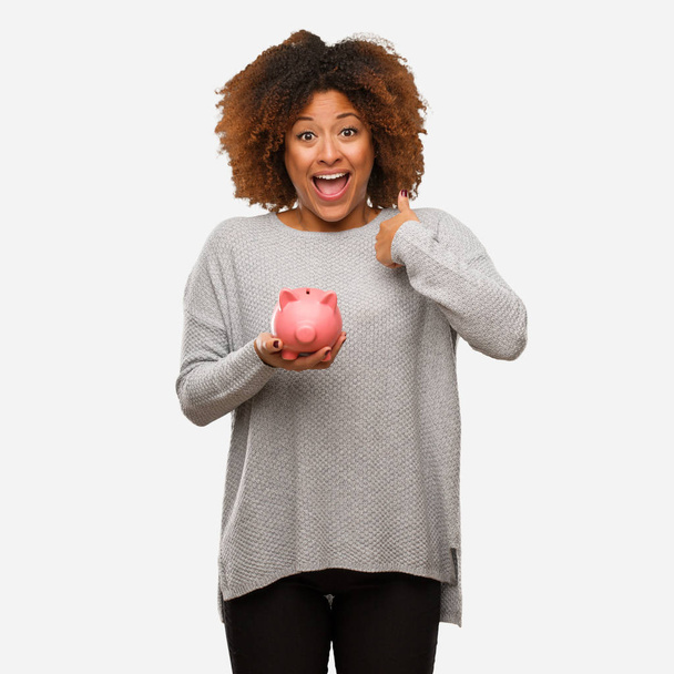 Young black woman holding piggy bank surprised, feels successful and prosperous - 写真・画像