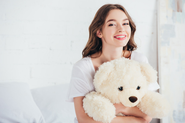 selective focus of happy woman smiling and hugging teddy bear in bed - Fotó, kép