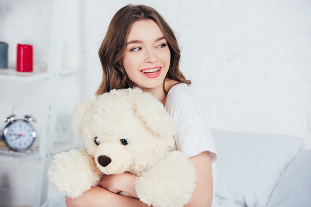 selective focus of smiling woman smiling and hugging teddy bear in bed - Foto, immagini