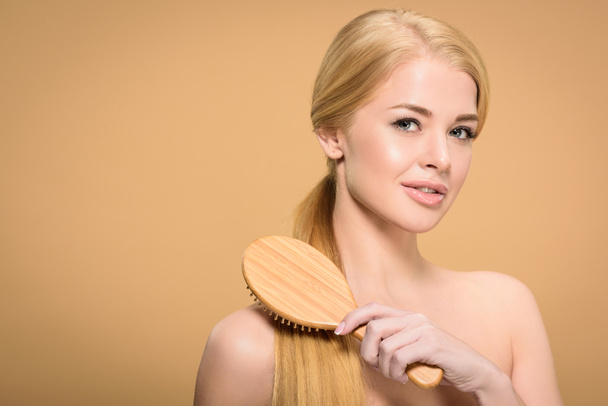 attractive blonde woman brushing smooth hair with wooden hairbrush and smiling at camera isolated on beige  - Photo, Image