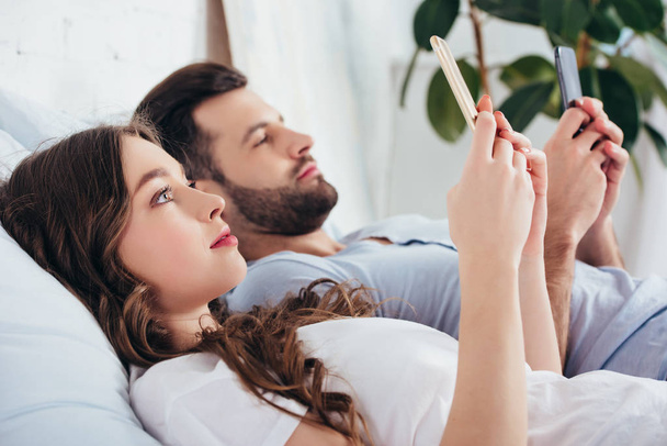 young couple relaxing in bed while using smartphones - Zdjęcie, obraz