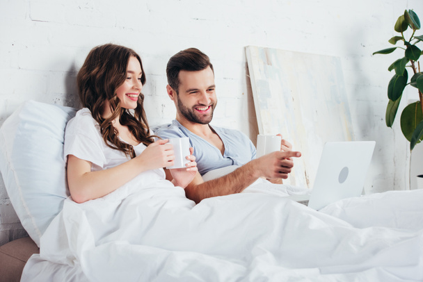 young couple holding cups, lying on soft white bedding and watching movie on laptop - Fotografie, Obrázek