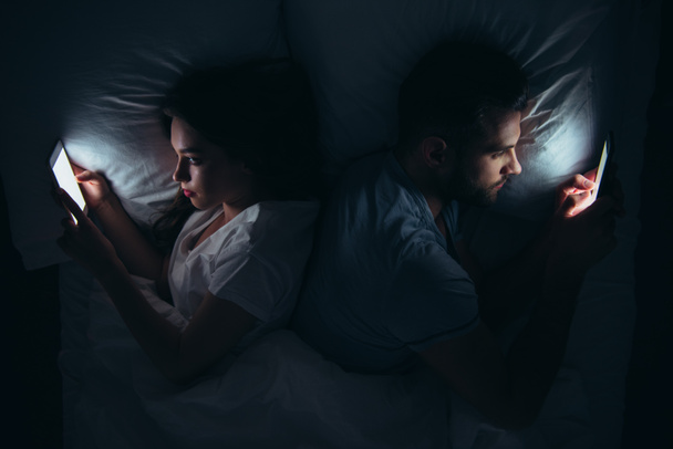couple lying back to back in bed and using smartphones at night - 写真・画像