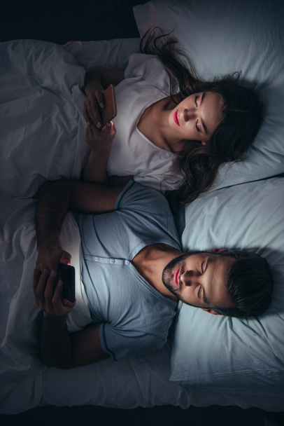 νεαρό ζευγάρι ξαπλωμένη στο κρεβάτι και χρησιμοποιώντας τα smartphones το βράδυ - Φωτογραφία, εικόνα