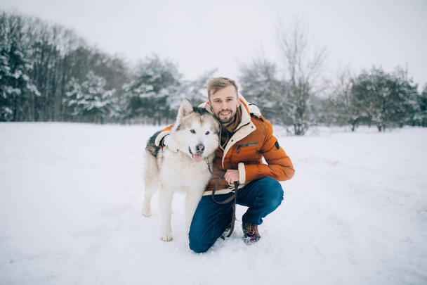 Młody człowiek siedzi w śniegu obok pies Alaskan Malamute i uściski jego na spacer zimą. - Zdjęcie, obraz
