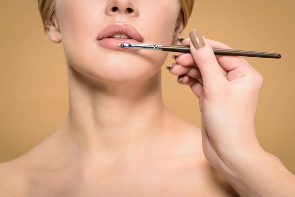 cropped shot of stylist with cosmetic brush applying lipstick to beautiful young woman isolated on beige - Photo, Image