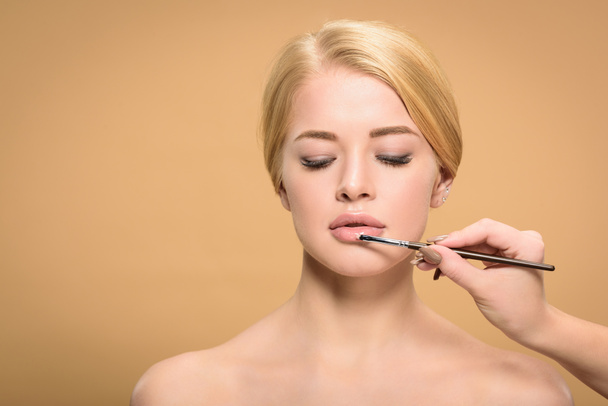 cropped shot of stylist holding cosmetic brush and applying lipstick to beautiful blonde girl isolated on beige - Foto, Imagem