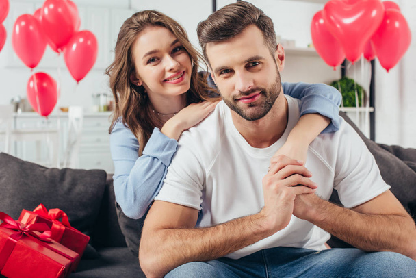 happy young girlfriend embracing boyfriend in room with heart-shaped balloons - Фото, изображение