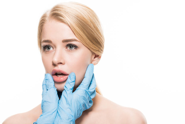περικοπεί shot από cosmetologist σε γάντια λατέξ, αγγίζοντας το πρόσωπο και τα χείλη της νεαρής γυναίκας που απομονώνονται σε λευκό - Φωτογραφία, εικόνα