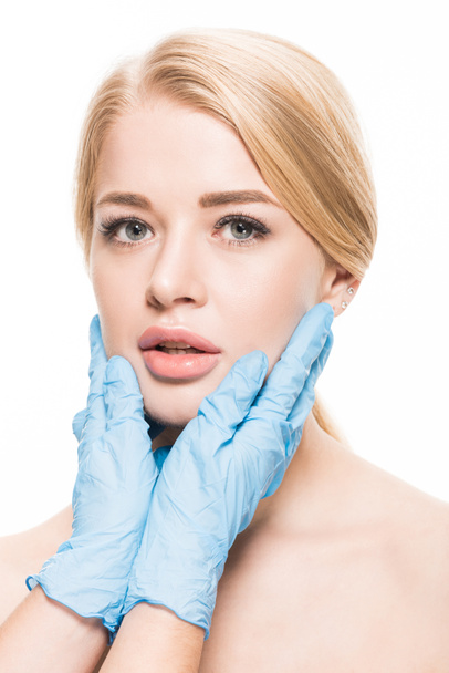 abgeschnittene Aufnahme eines Kosmetikers in medizinischen Handschuhen, der das Gesicht einer jungen Frau berührt, die isoliert auf Weiß in die Kamera blickt  - Foto, Bild