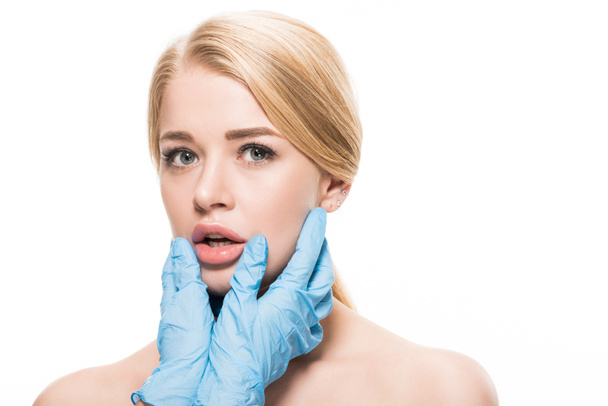 cropped shot of cosmetologist in latex gloves holding chin of young woman looking at camera isolated on white  - Photo, Image