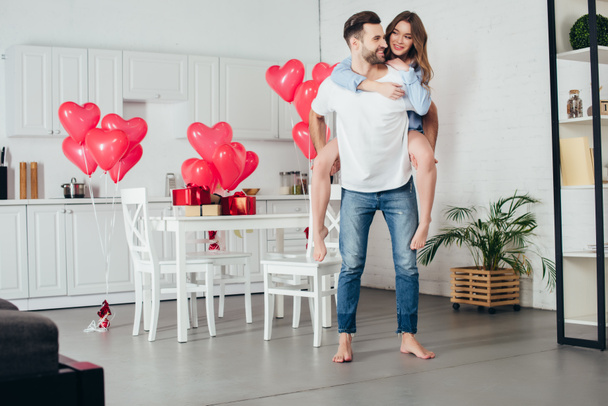 glückliches Paar huckepack zu Hause in möbliertem Zimmer mit herzförmigen Luftballons auf dem Hintergrund - Foto, Bild