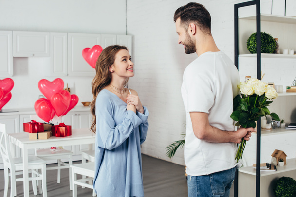 young smiling girl in joyful expectation of st valentines day gift while smiling boyfriend holding roses bouquet behind back  - Photo, Image