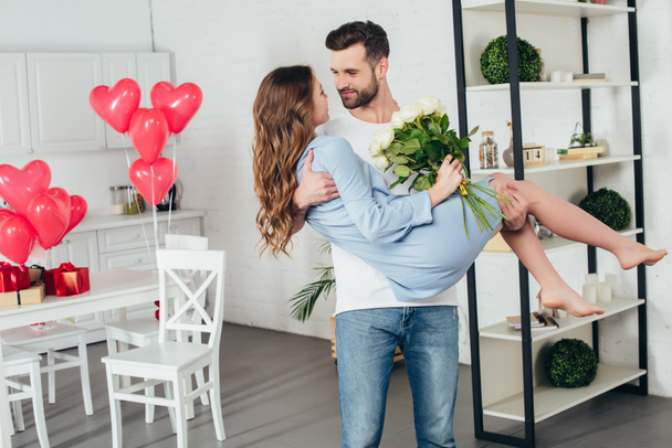 young man holding smiling girlfriend with roses bouquet in arms at st valentine day - Фото, изображение