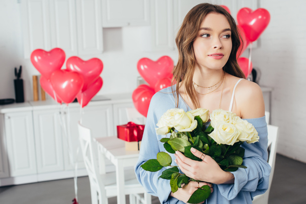 selective focus of a smiling girl holding bouquet of roses in room decorated with heart-shaped balloons  - Photo, Image