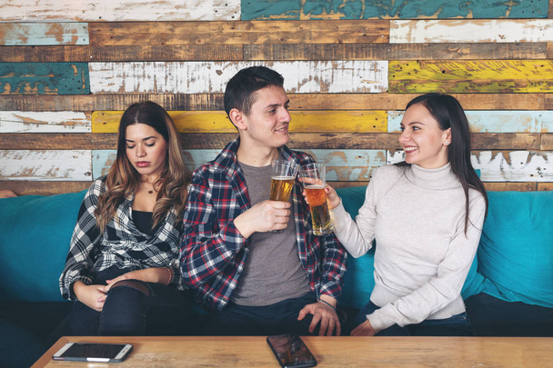 Happy young girl drinking beer with young man and socialise ignoring other jealous sad woman sitting next to them at rustic bar restaurant. Love and jealousy concept of young generation of teens - Fotoğraf, Görsel
