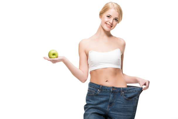 beautiful slim girl in oversized jeans holding fresh apple and smiling at camera isolated on white - Fotografie, Obrázek