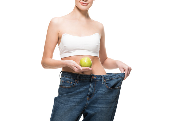 cropped shot of smiling young woman in oversized jeans holding green apple isolated on white  - Foto, afbeelding