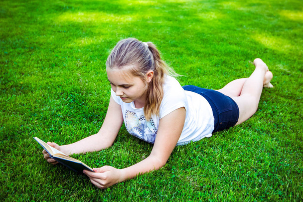 Beautiful teenage girl lies on green grass and reads book in park on summer sunny day. - Photo, Image