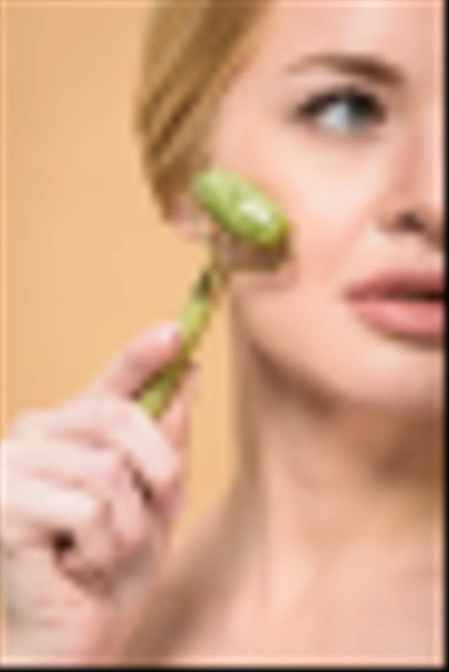 cropped shot of young woman massaging face with jade roller and looking away isolated on beige - 写真・画像