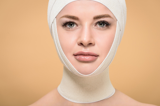 молодая женщина с бинтами над головой, смотрящая на камеру, изолированную от бежевого, концепция пластической хирургии
 - Фото, изображение