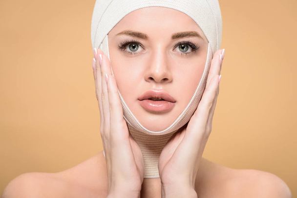 young woman with bandages over head touching face and looking at camera isolated on beige - Foto, Imagen
