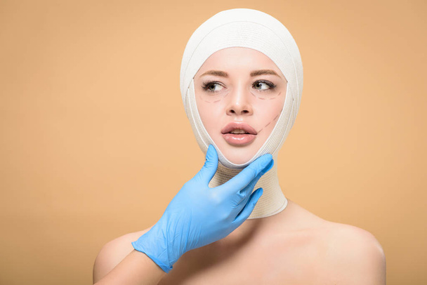 doctor in latex glove touching face of young woman with bandages on head and correcting lines isolated on beige  - Foto, afbeelding