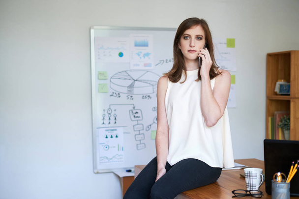 Молодая красивая деловая женщина сидит на деревянном рабочем столе в офисе и имеет телефонный звонок глядя в камеру
 - Фото, изображение