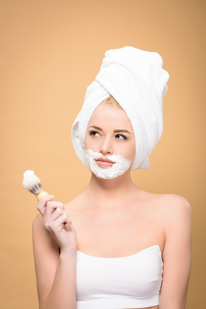 woman with towel on head and shaving cream on face holding shaving brush and looking away isolated on beige - Photo, image