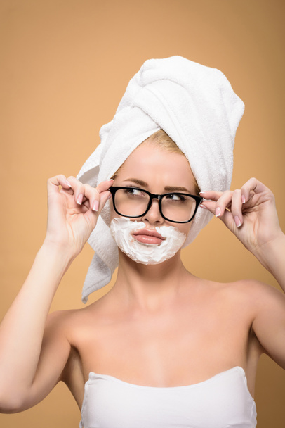 beautiful girl with towel on head and shaving cream on face adjusting eyeglasses and looking away isolated on beige - Fotó, kép