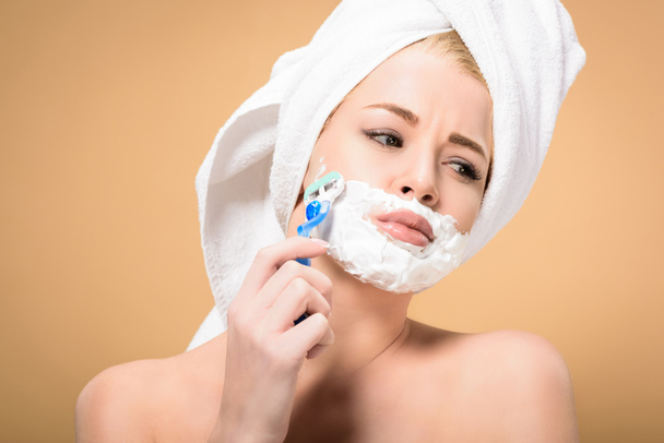 mujer joven desnuda con toalla en la cabeza afeitándose la cara con afeitadora y mirando hacia otro lado aislado en beige
 - Foto, imagen
