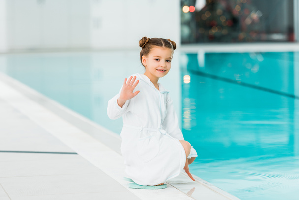 Niedliches Kind sitzt im Bademantel im Schwimmbad und winkt freundlich  - Foto, Bild
