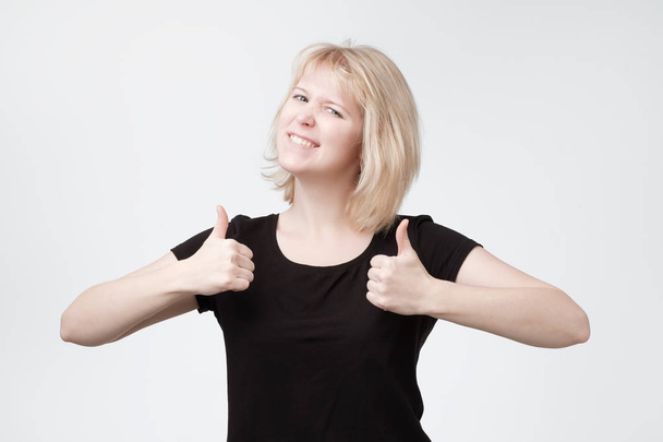 Lachende mooie jonge blonde tiener duimen opdagen. Europese vrouw met positieve stemming geïsoleerd op de witte achtergrond - Foto, afbeelding