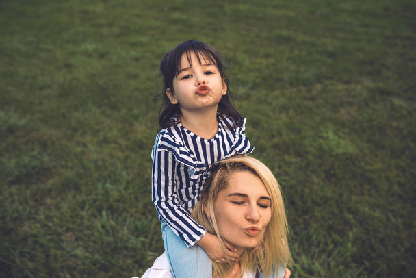 Image of beautiful woman playing and blowing kiss with her cute little girl on green grass outdoor. Loving pretty mother and daughter spend time together in the park. Mom and kid have fun.Happy family - Φωτογραφία, εικόνα