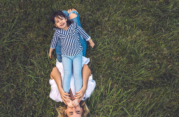 Overhead view of beautiful woman playing with her cute little girl on green grass outdoor. Loving happy pretty mother and happy daughter spend time together in park. Motherhood, childhood. Copy space - Photo, image